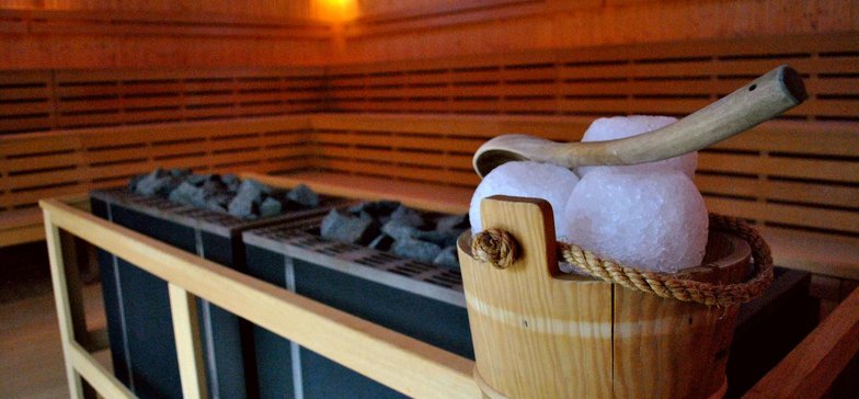 Piece w dużej saunie zewnętrznej. Na drewnianej obudowie ustawione drewniane wiaderko na wodę, na którym leżą trzy lodowe kule i łyżka do rozlewania wody.