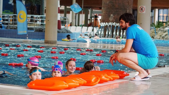 Basen sportowy wewnętrzny. Cztery dziewczynki w wodzie trzymają się krawędzi basenu słuchając wskazówek instruktora w niebieskim uniformie. Na brzegu leży kilka pomarańczowych bojek ratowniczych. W tle pływają inne dzieci. 