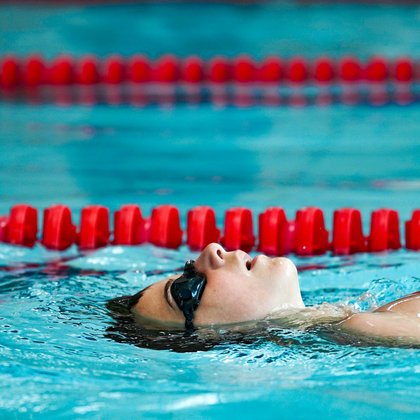 Zbliżenie na chłopca pływającego w basenie sportowym, ma na sobie ciemne okularki pływackie, w tle widać liny rozdzielające kolejne tory. 