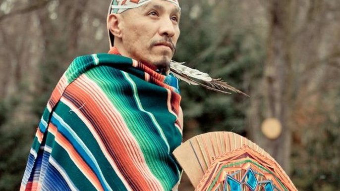 Mężczyzna w średnim wieku o południowej urodzie ma na sobie kolorowy strój inspirowany kulturą Indian - w dłoniach trzyma kolorowy wachlarz, na głowie ma opaskę zakończoną piórami. 