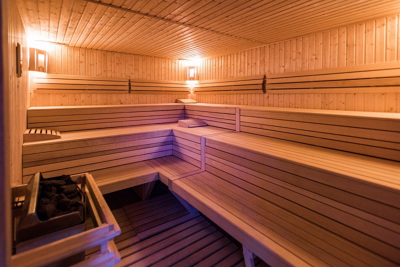 Wnętrze wewnętrznej sauny fińskiej, po lewej stronie przy drzwiach piec z ciemnymi kamieniami, naprzeciw i po prawej stronie trzy rzędy siedzisk. Cała sauna zabudowana drewnem.